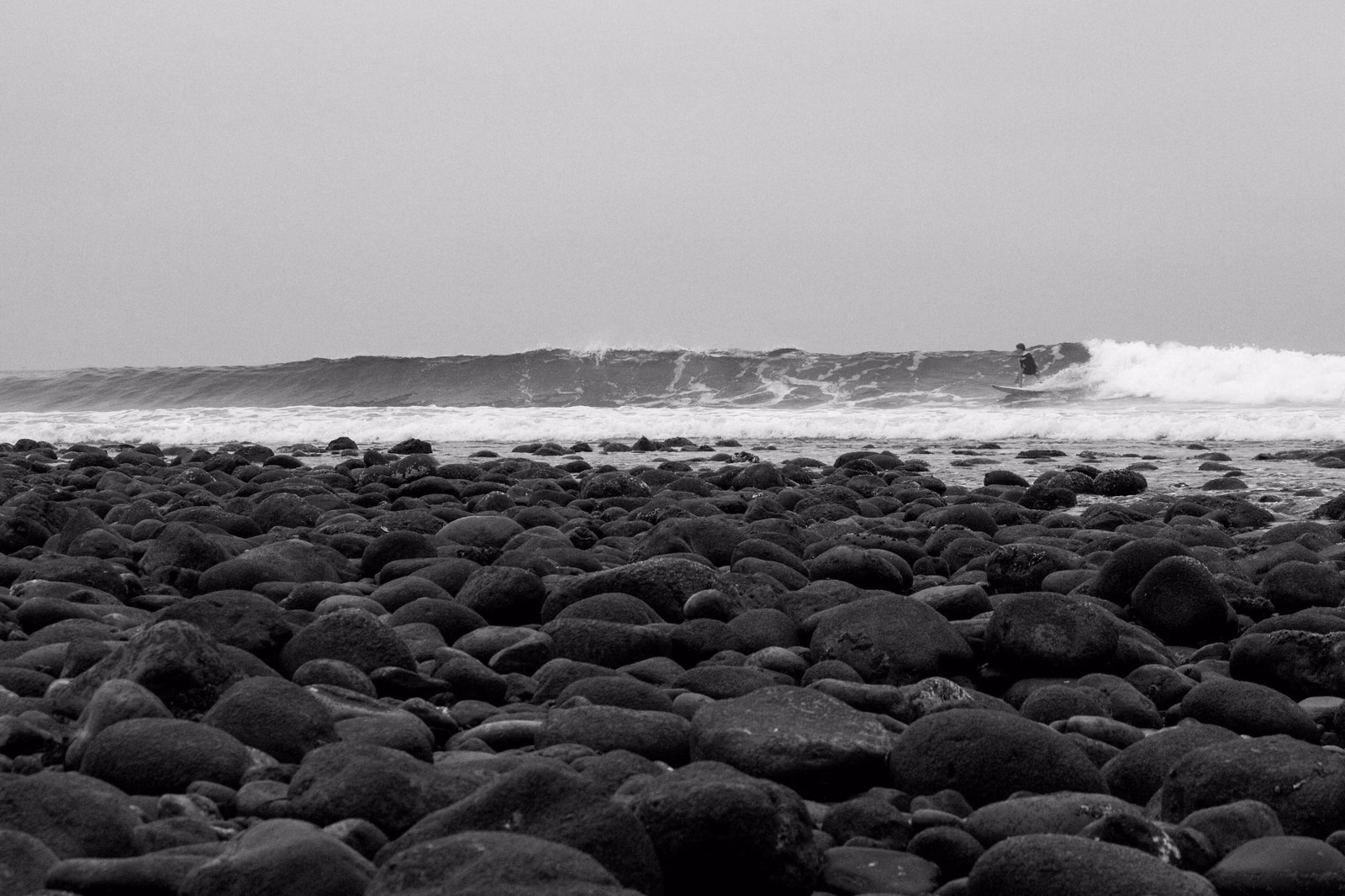 Cobblestone beach with a surfer riding a long wave in the distance
